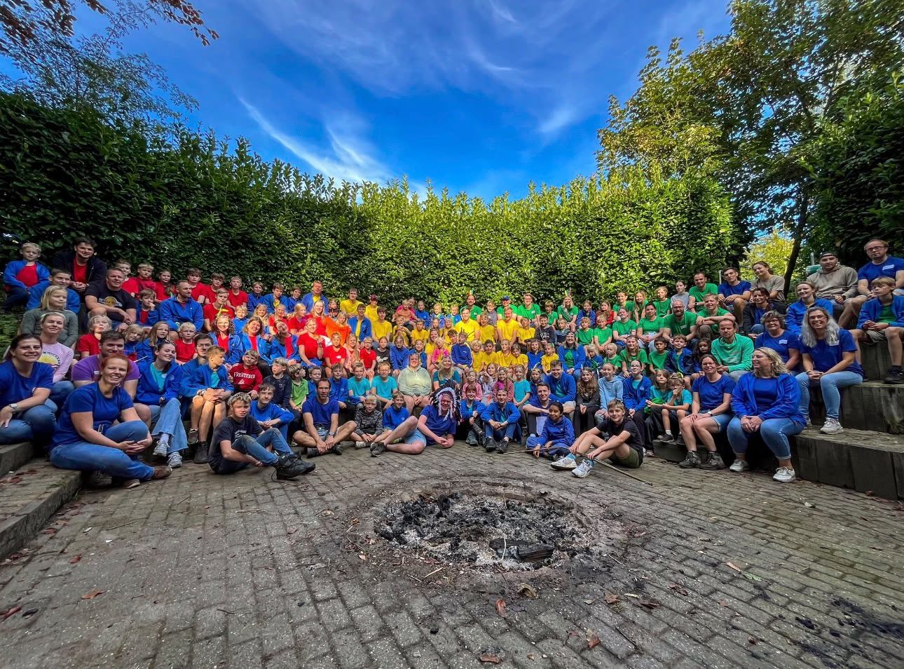 Alle leden en leiding van Scouting Groenlo zitten samen op de treden van de theaterkuil, rond de kampvuurplaats.
